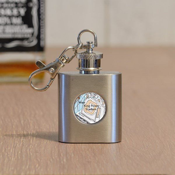 A stainless steel flask keyring with a silver metal clasp, resting on a wooden surface. The front of the flask features a circular clear cover displaying a colourful map design with the name 'King Power Stadium' written on it.