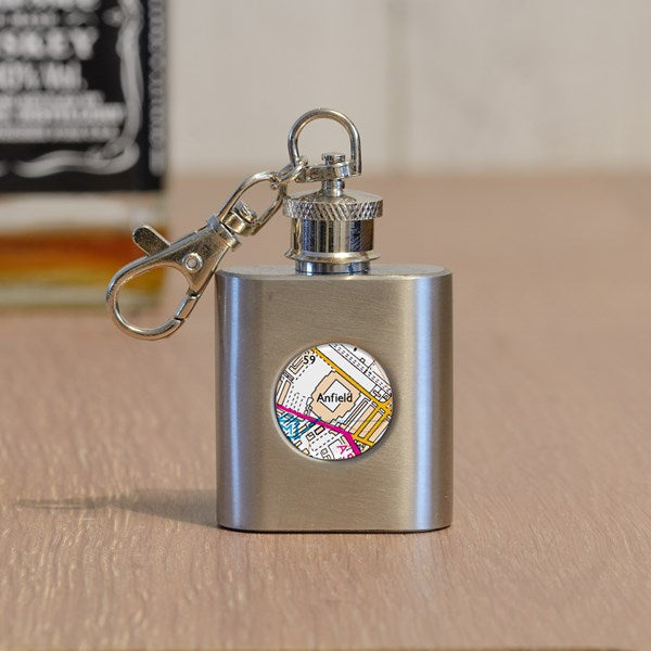 A stainless steel flask keyring with a silver metal clasp, resting on a wooden surface. The front of the flask features a circular clear cover displaying a colourful map design with the name 'Anfield' written on it.