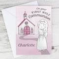 A personalized First Holy Communion greeting card with a pink background, featuring an illustration of a child in a white dress next to a pink church, with the name 'Charlotte' printed at the bottom. The card is shown with a white envelope on a light gray wood-textured background.