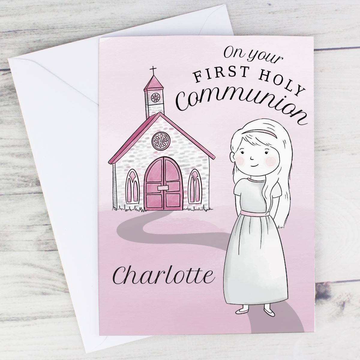 A personalized First Holy Communion greeting card with a pink background, featuring an illustration of a child in a white dress next to a pink church, with the name 'Charlotte' printed at the bottom. The card is shown with a white envelope on a light gray wood-textured background.