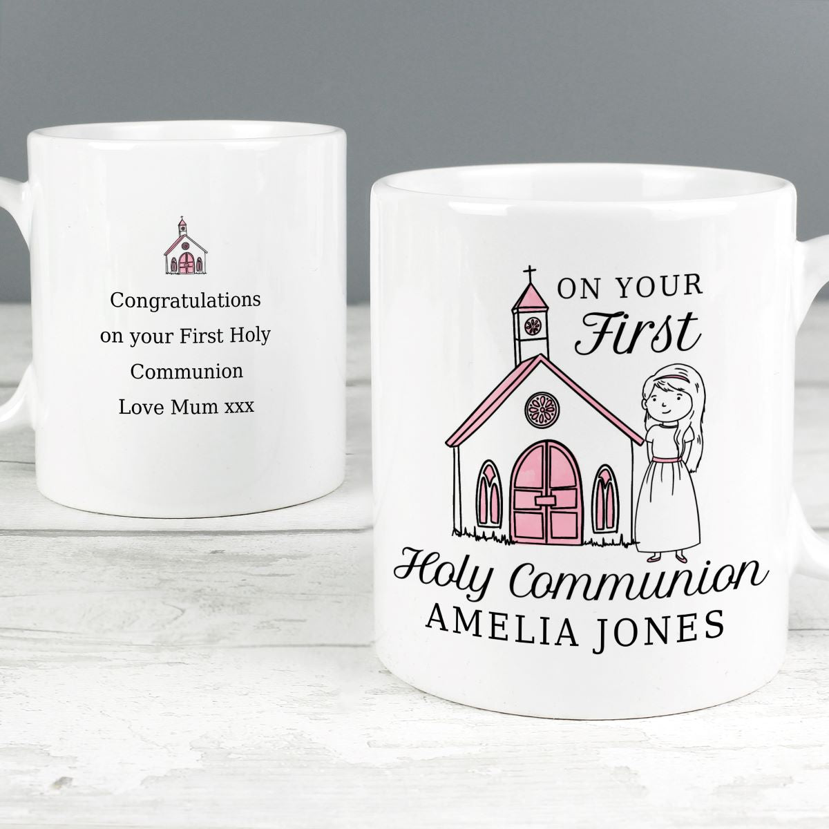 Two white ceramic mugs shown on a light wood surface against a gray background. One mug is turned away and shows black text reading 'Congratulations on your First Holy Communion Love Mum xxx' with a small church illustration.