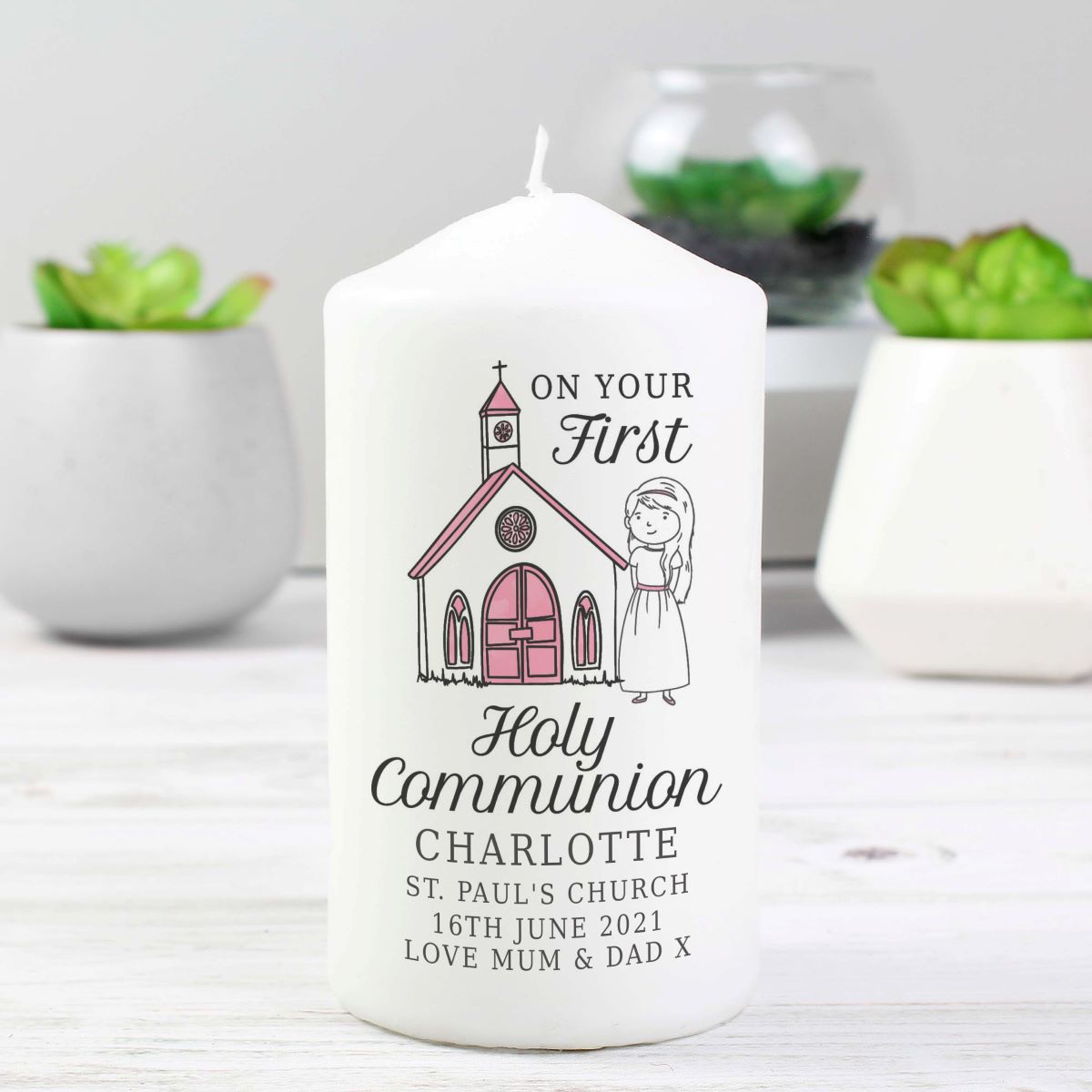 White candle with a printed design of a church and text, placed on a white surface with plants in the background.