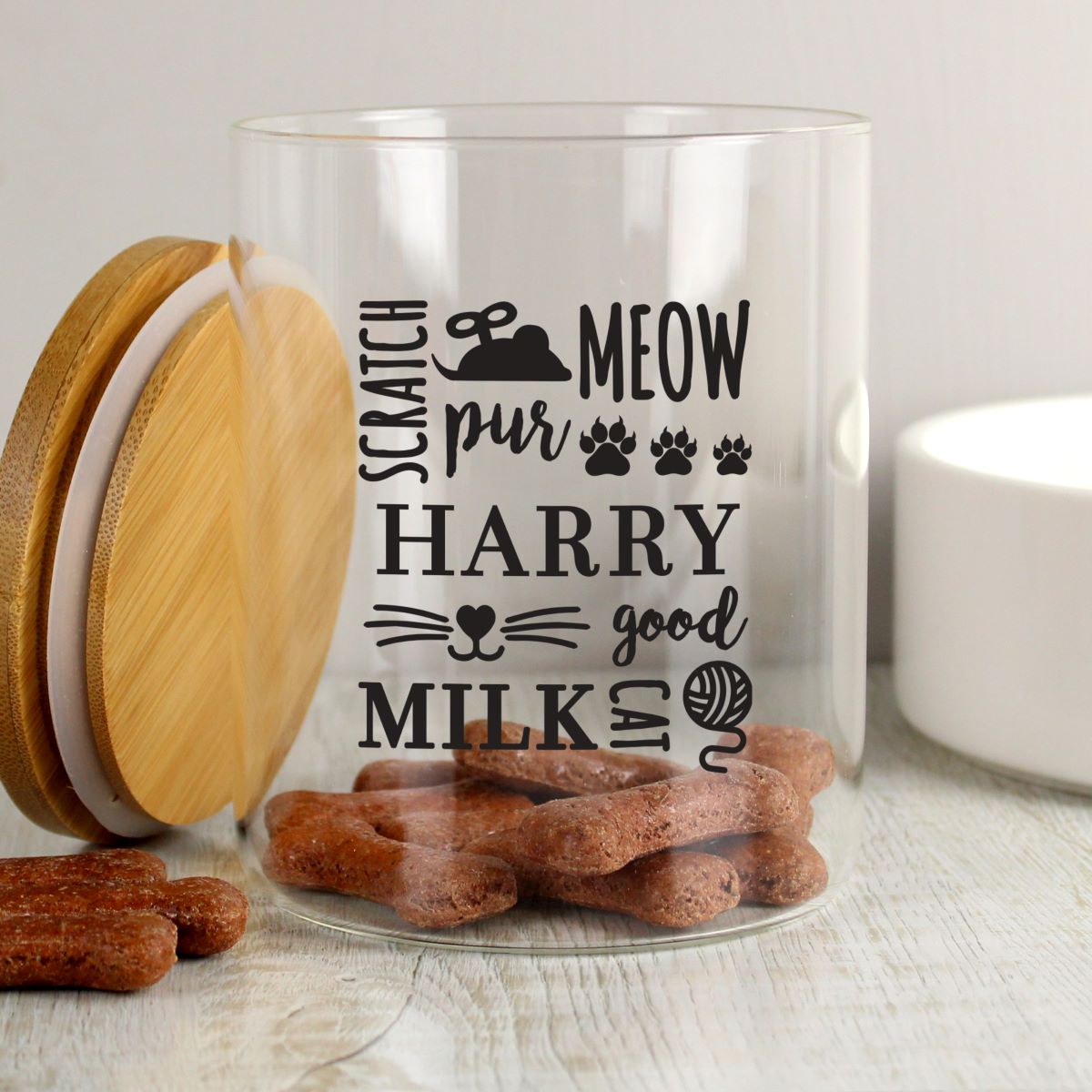 A clear cylindrical glass jar with a bamboo lid, filled with brown treats and printed with the text 'SCRATCH pur MEOW HARRY good MILKS' with cat and dog-themed graphics. The jar is set on a light wooden surface with a blurred white background.