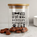 Clear pet treat jar with wooden lid, personalized text, and dog treats on a light surface.