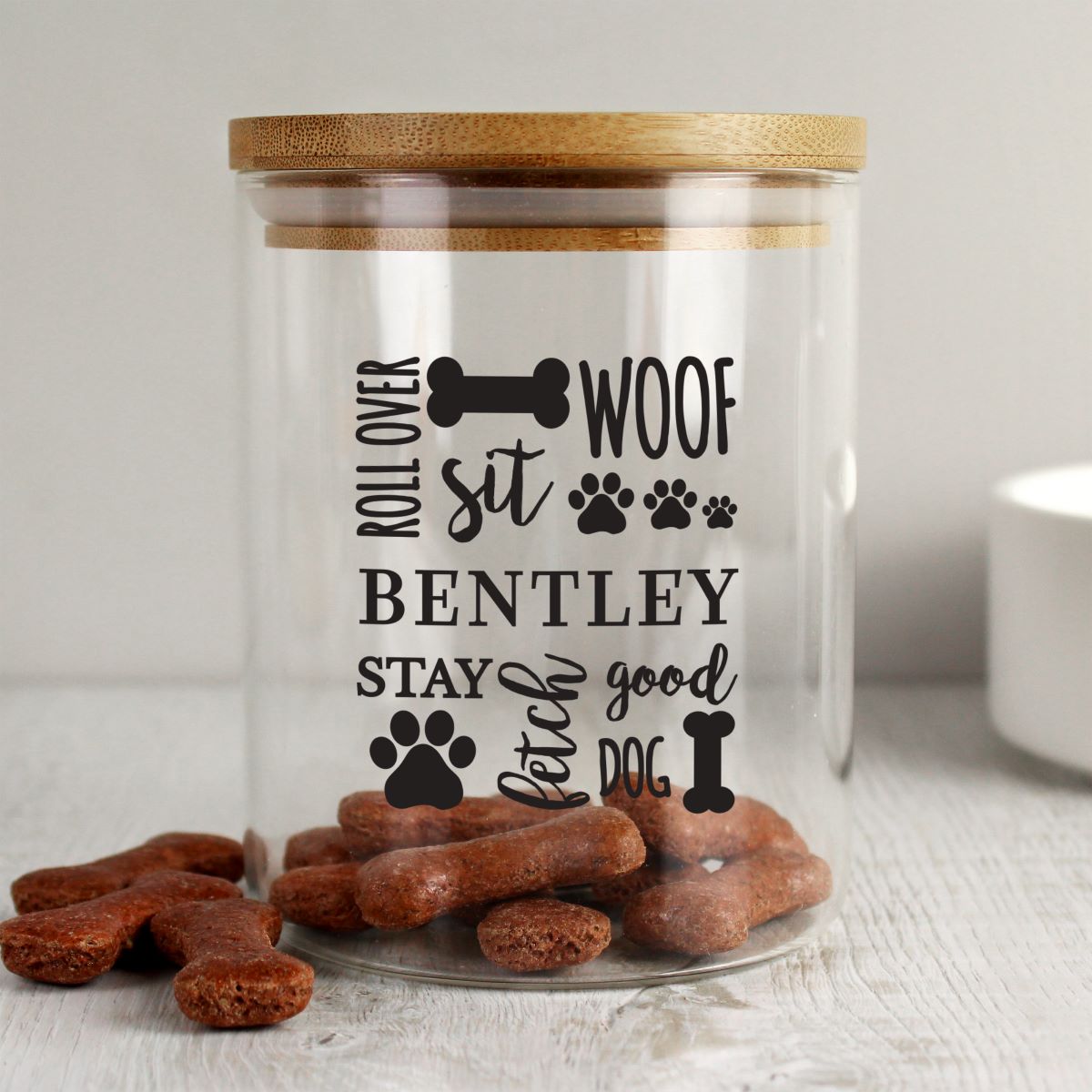 Clear pet treat jar with wooden lid, personalized text, and dog treats on a light surface.