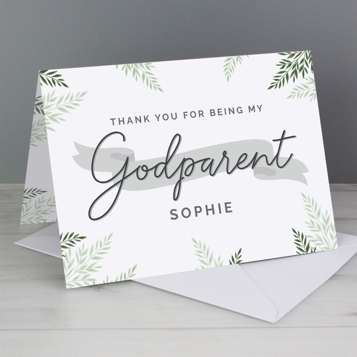 A personalized thank you card for a godparent, displayed standing on a white envelope against a gray background. The card features green leaf illustrations in the corners, a gray brushstroke design, and the text 'THANK YOU FOR BEING MY Godparent SOPHIE'.