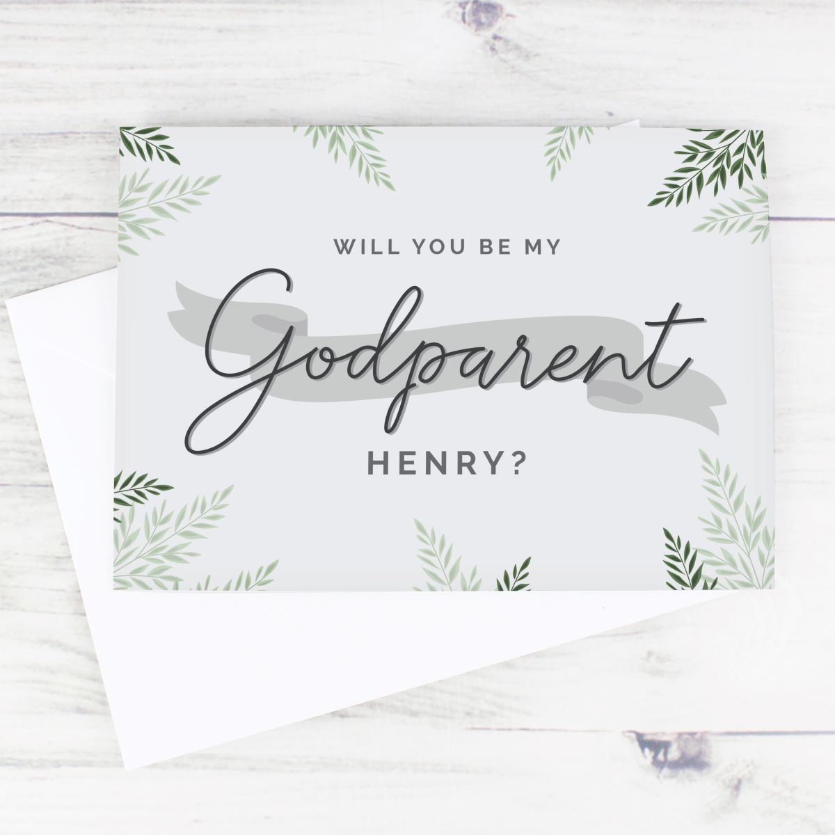 A square wedding card with a white envelope, featuring the text 'WILL YOU BE MY Godparent HENRY?' in black script and sans-serif fonts, decorated with green leaf illustrations on a light gray background, set on a white wood-textured surface.
