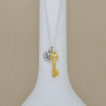 A silver chain necklace with a gold key pendant and a silver disc charm engraved with the letter 'M', displayed on a white neck stand against a beige background.