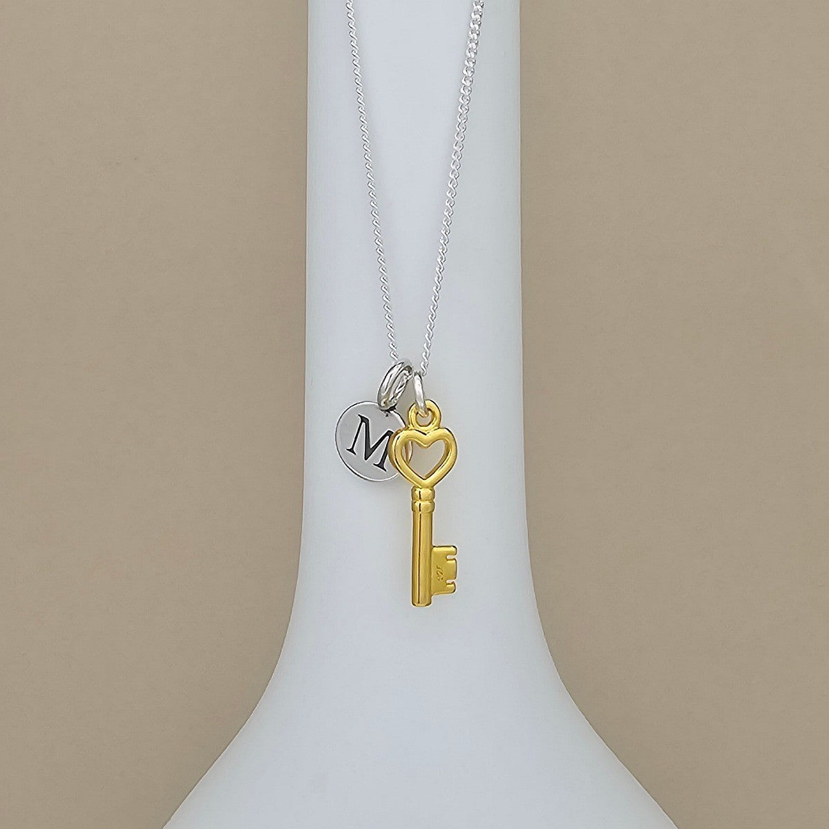 A silver chain necklace with a gold key pendant and a silver disc charm engraved with the letter 'M', displayed on a white neck stand against a beige background.