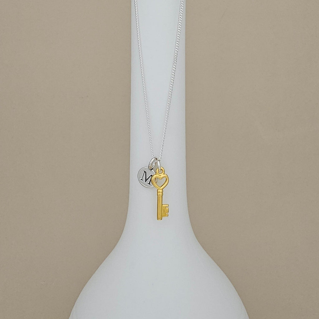 A silver chain necklace with a gold key charm and a small silver heart charm engraved with the letter 'M', displayed on a white vase against a beige background.