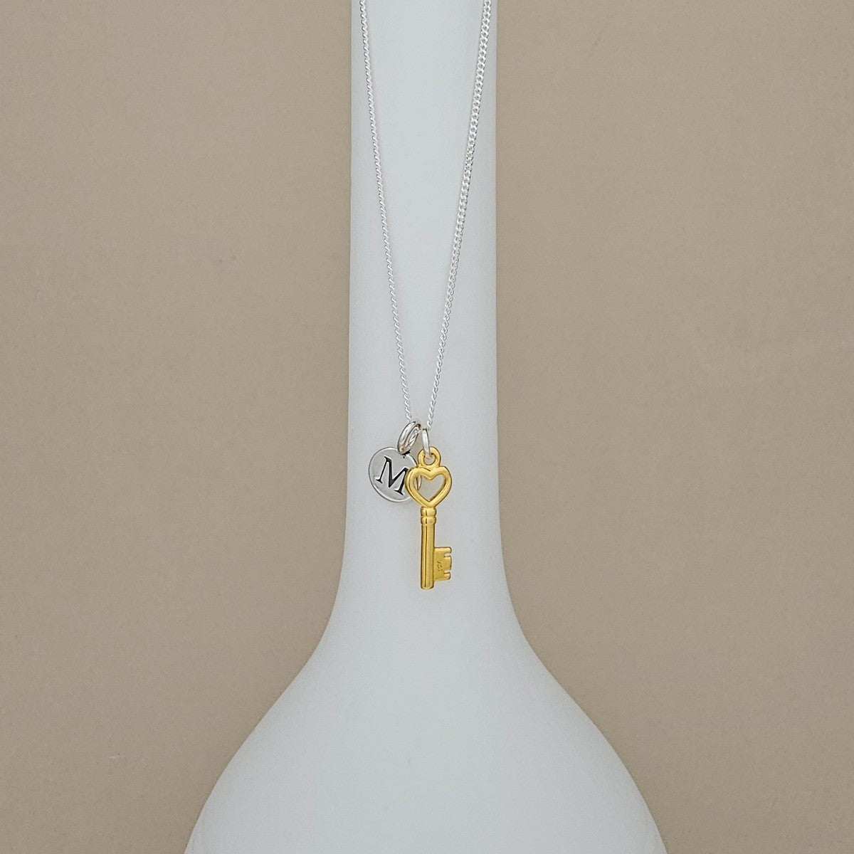A silver chain necklace with a gold key charm and a small silver heart charm engraved with the letter 'M', displayed on a white vase against a beige background.