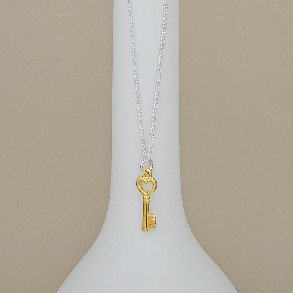 A gold key pendant necklace with a heart-shaped key head hangs from a delicate silver chain, displayed on a white ceramic vase against a warm beige background.