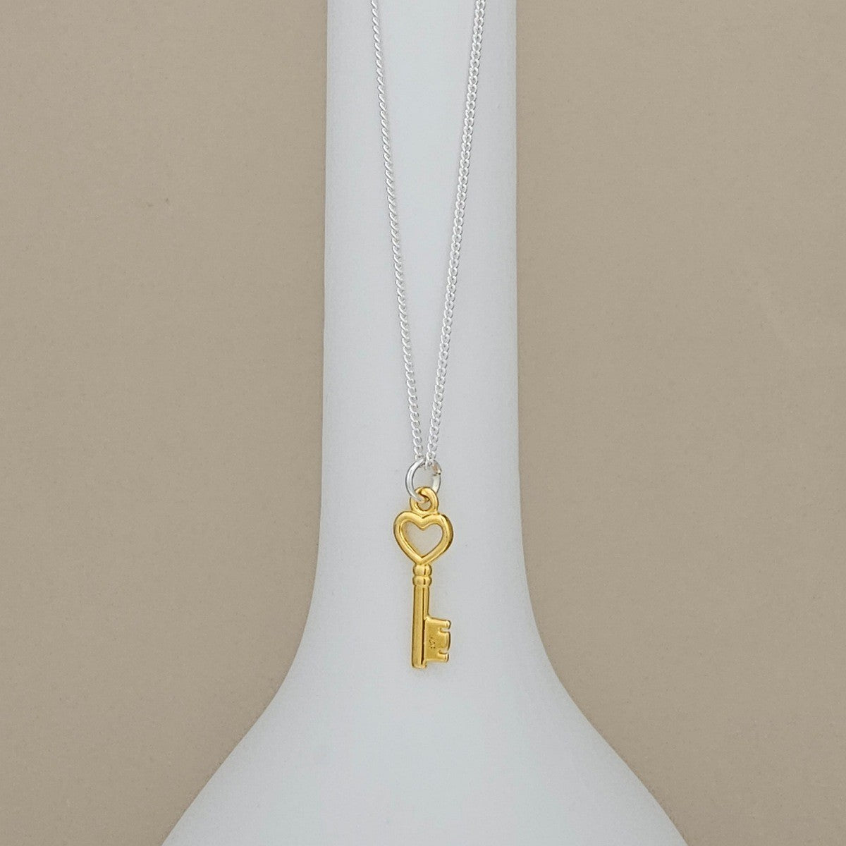 A gold key pendant necklace with a heart-shaped key head hangs from a delicate silver chain, displayed on a white ceramic vase against a warm beige background.