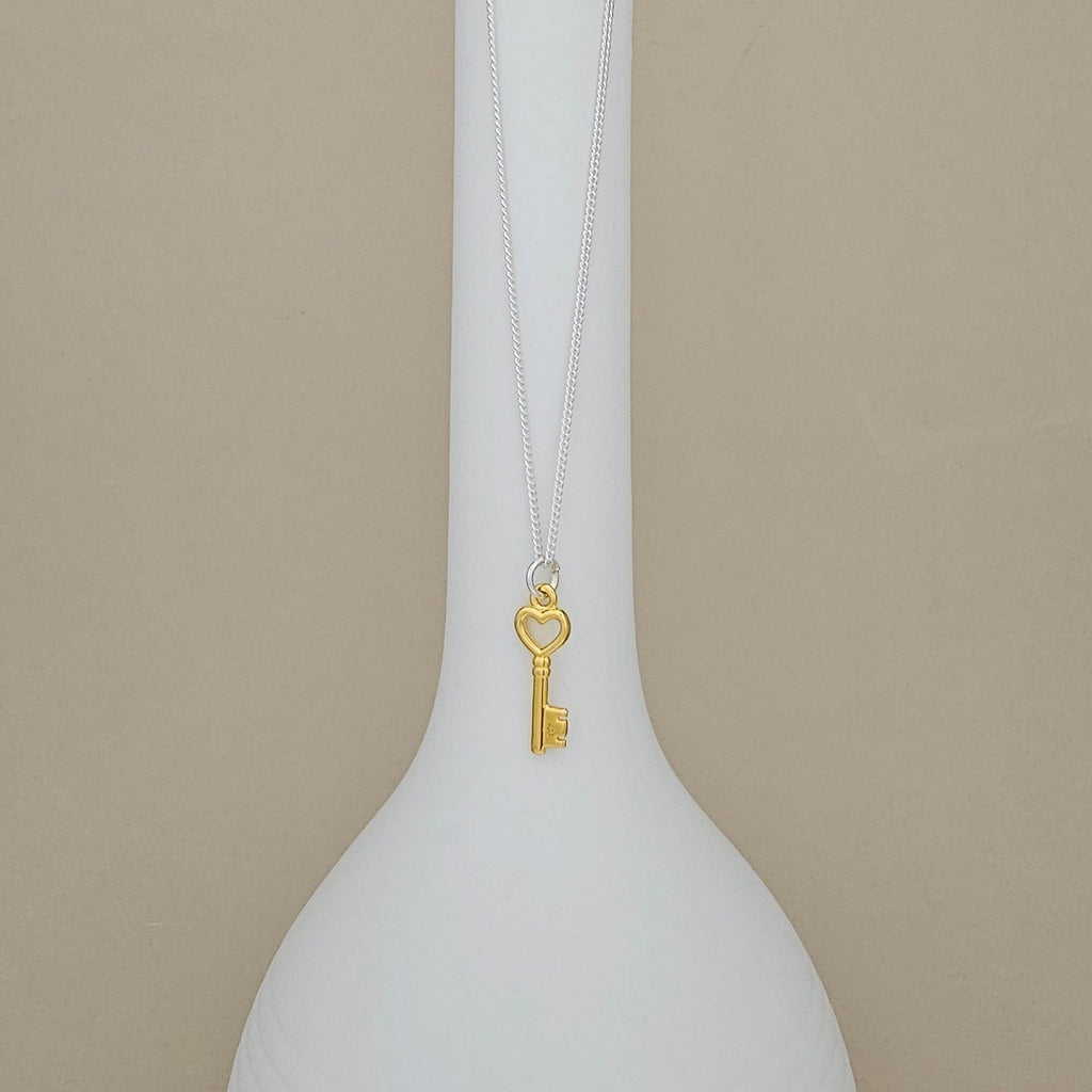 A gold key pendant necklace with a heart-shaped keyhole, hanging from a delicate silver chain on a white vase against a beige background.