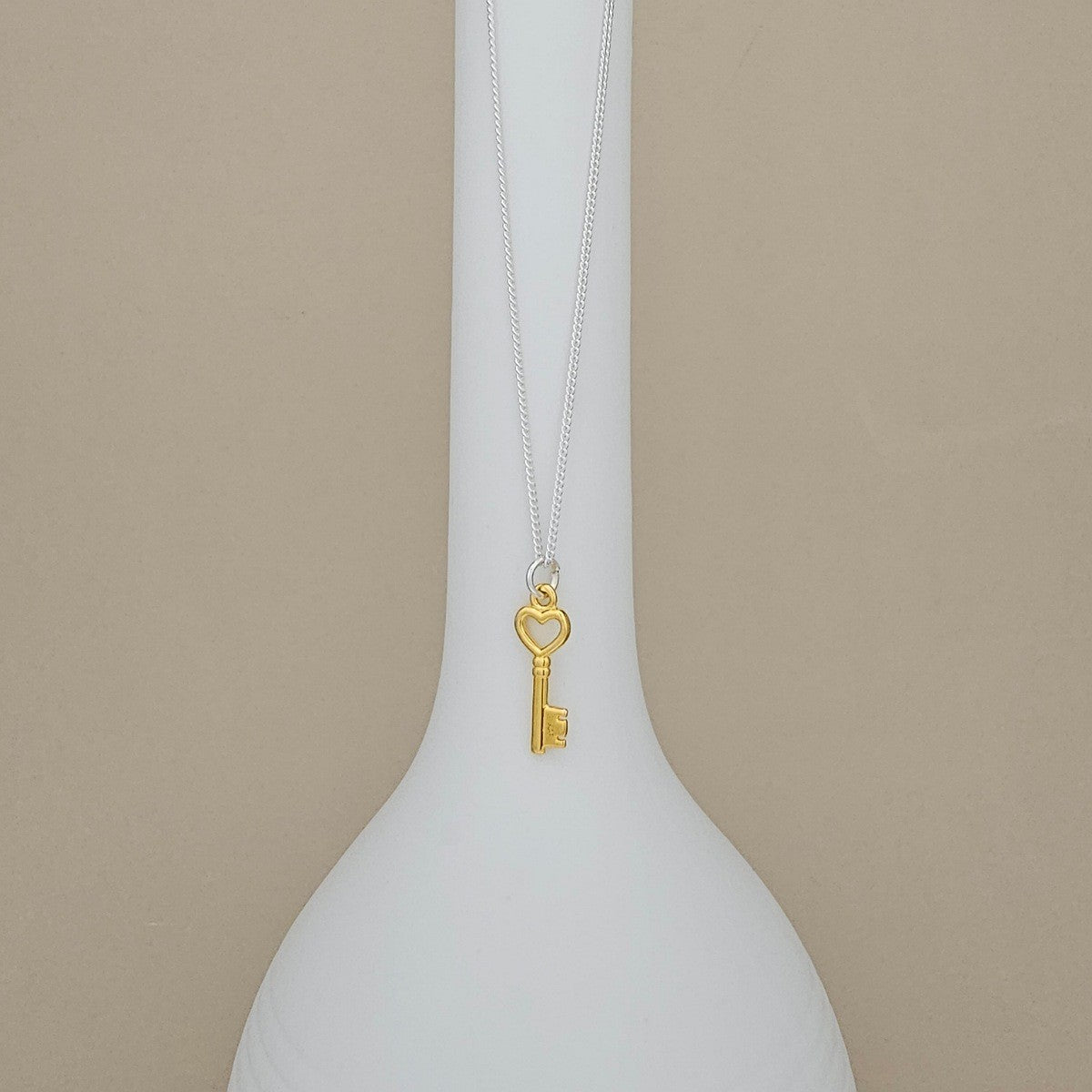 A gold key pendant necklace with a heart-shaped keyhole, hanging from a delicate silver chain on a white vase against a beige background.