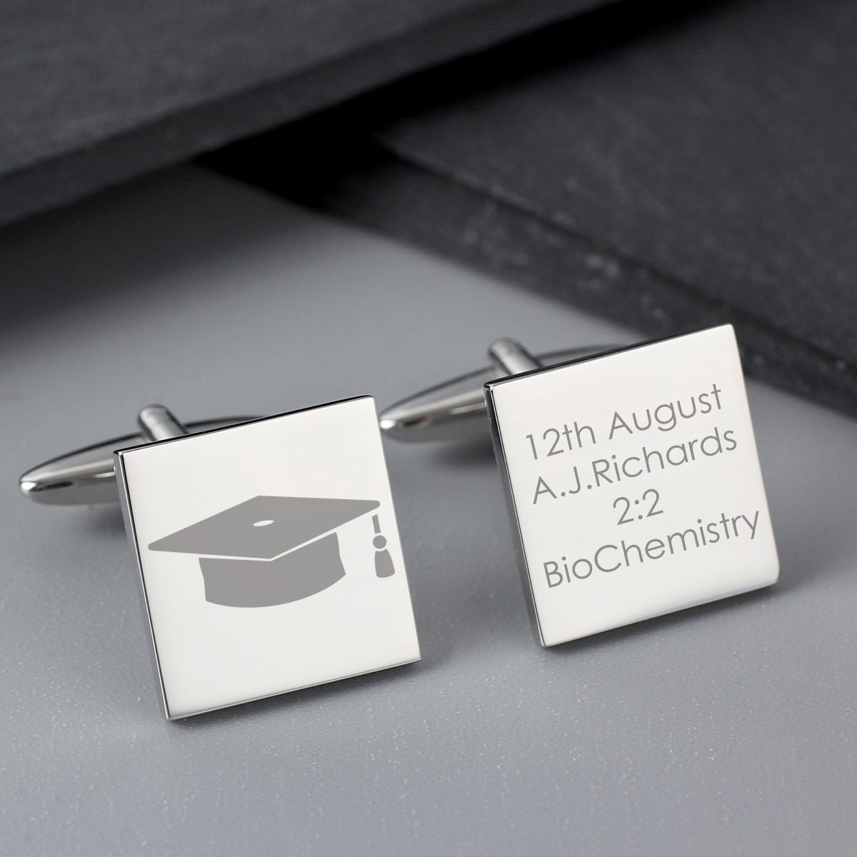 Silver cufflinks with graduation cap icon and engraved text on a gray surface