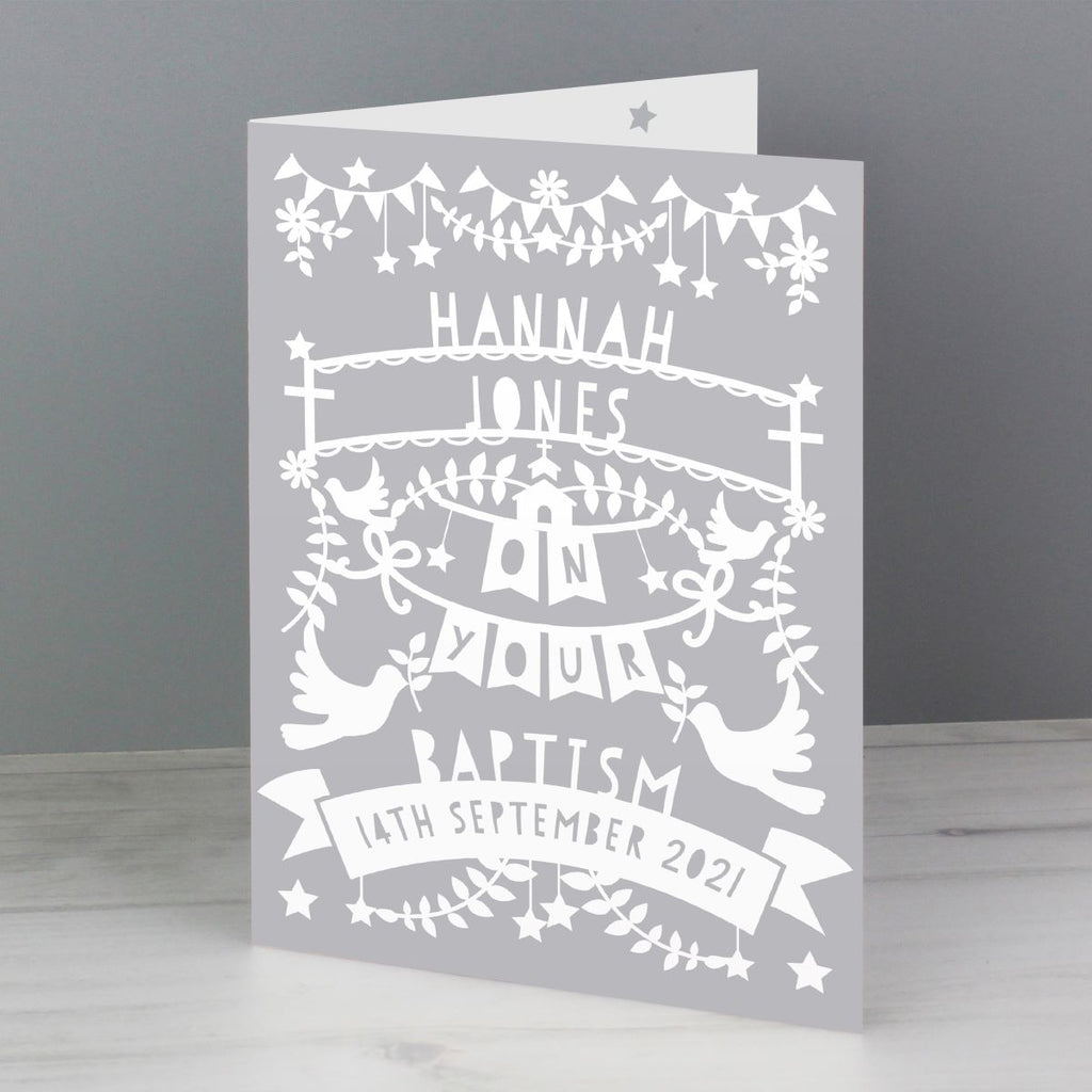 A folded greeting card standing upright on a light surface against a grey background. The card is light grey with white printed decorative illustrations and personalized text reading 'Hannah Jones on your baptism 14th September 2021'.