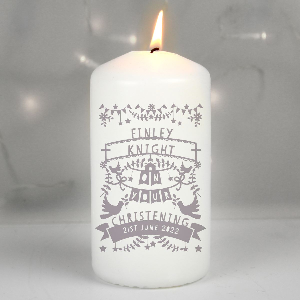 A white pillar candle with a lit flame, featuring a grey printed christening design with the text 'FINLEY KNIGHT ON YOUR CHRISTENING 21ST JUNE 2022'.
