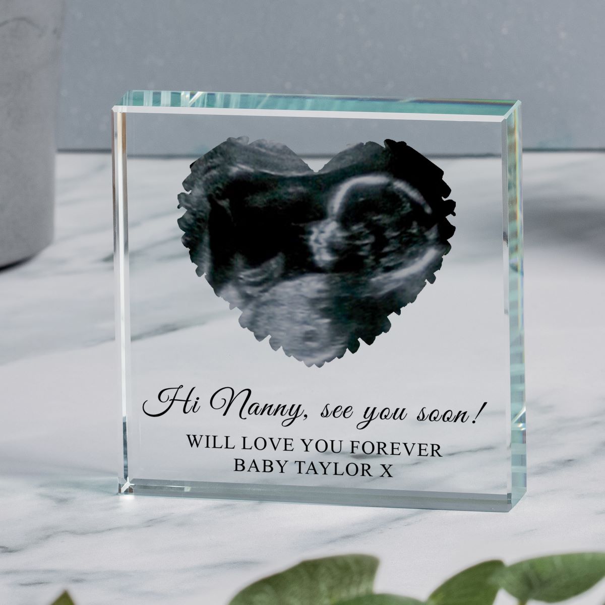 Clear glass block with a custom black and white ultrasound image in the shape of a heart and personalised text 'Hi nanny, see you soon! WILL LOVE YOU FOREVER BABY TAYLOR X' displayed on a Mable surface.