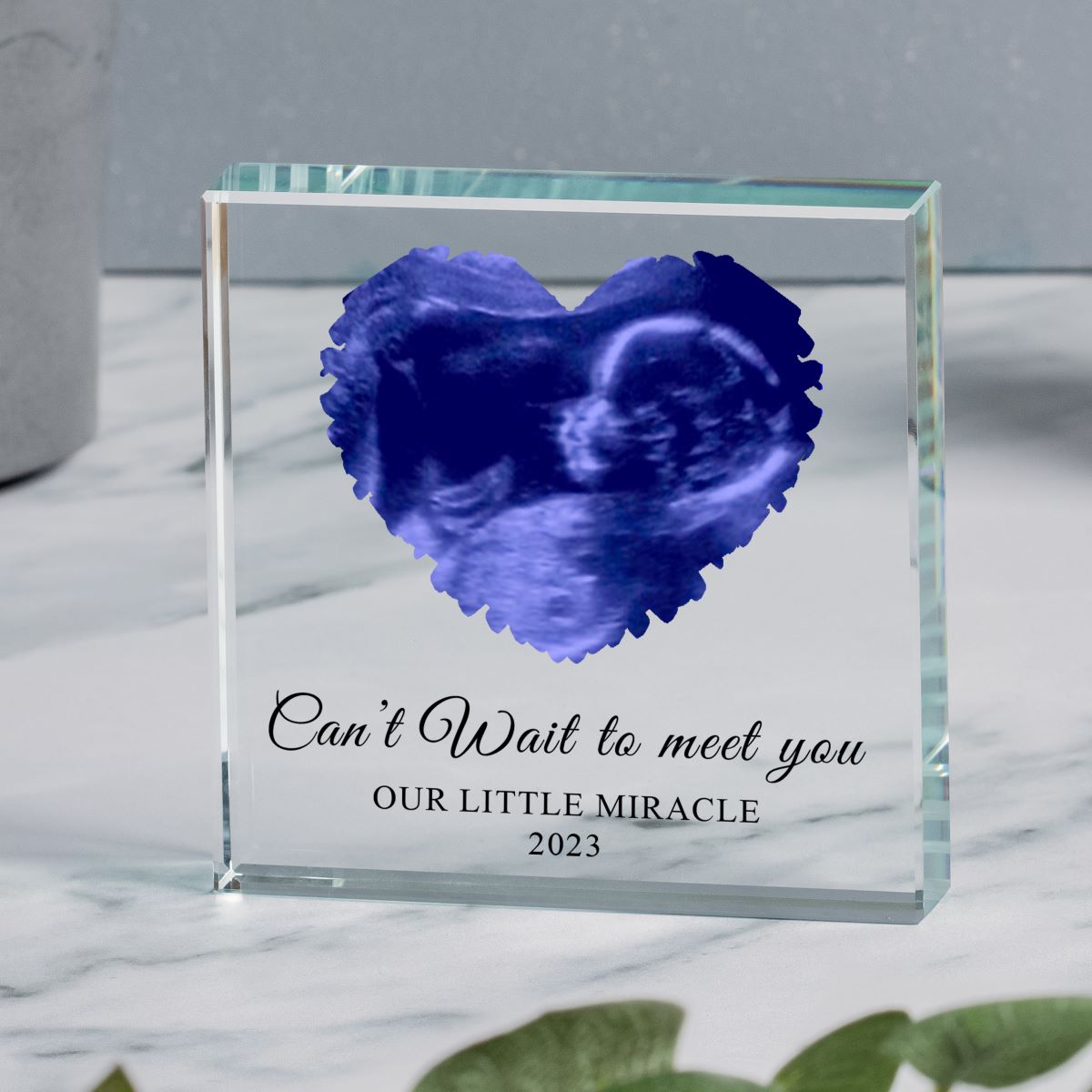 Clear glass block with a custom blue ultrasound image in the shape of a heart and personalised text 'Can't Wait to meet you, OUR LITTLE MIRACLE 2023' displayed on a Mable surface.