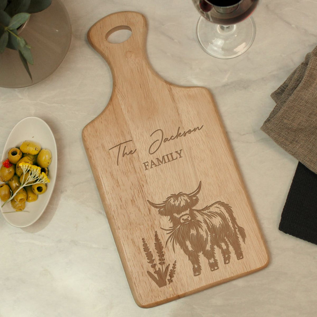 A personalized wooden cutting board with a handle, engraved with 'The Jackson FAMILY' and a Highland cow illustration, displayed on a light marble surface with styled food and tableware props.