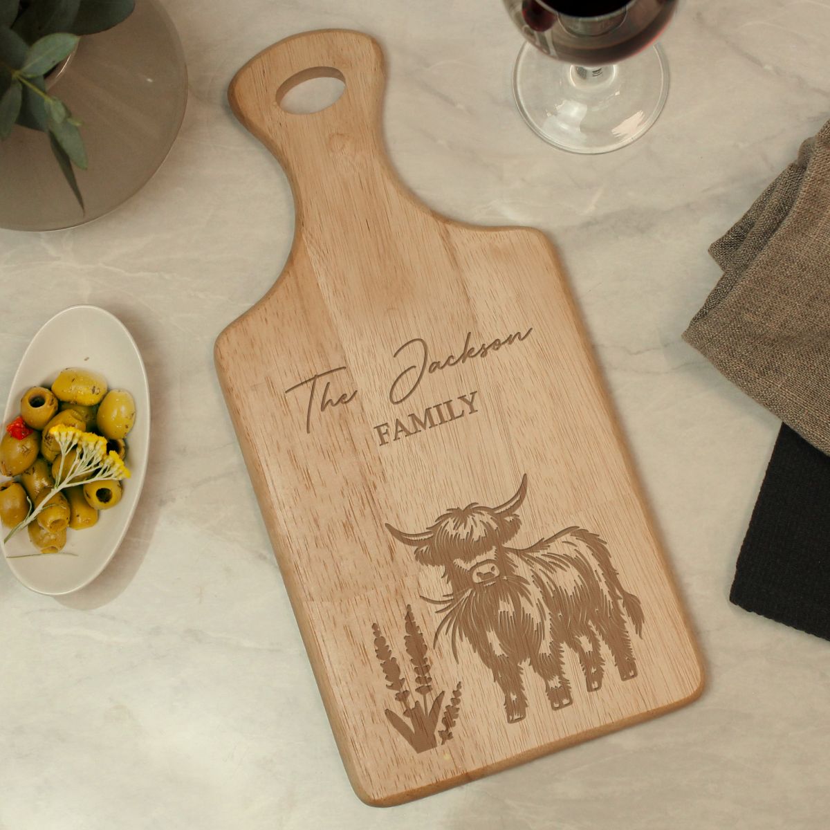 A personalized wooden cutting board with a handle, engraved with 'The Jackson FAMILY' and a Highland cow illustration, displayed on a light marble surface with styled food and tableware props.