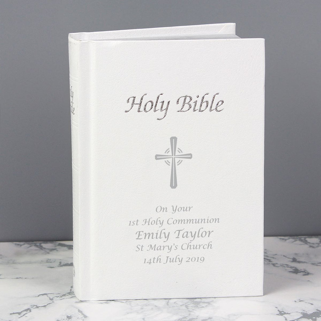 A personalized Holy Bible with a white cover, featuring a silver cross and custom text for a 1st Holy Communion, displayed on a marble surface against a gray background.
