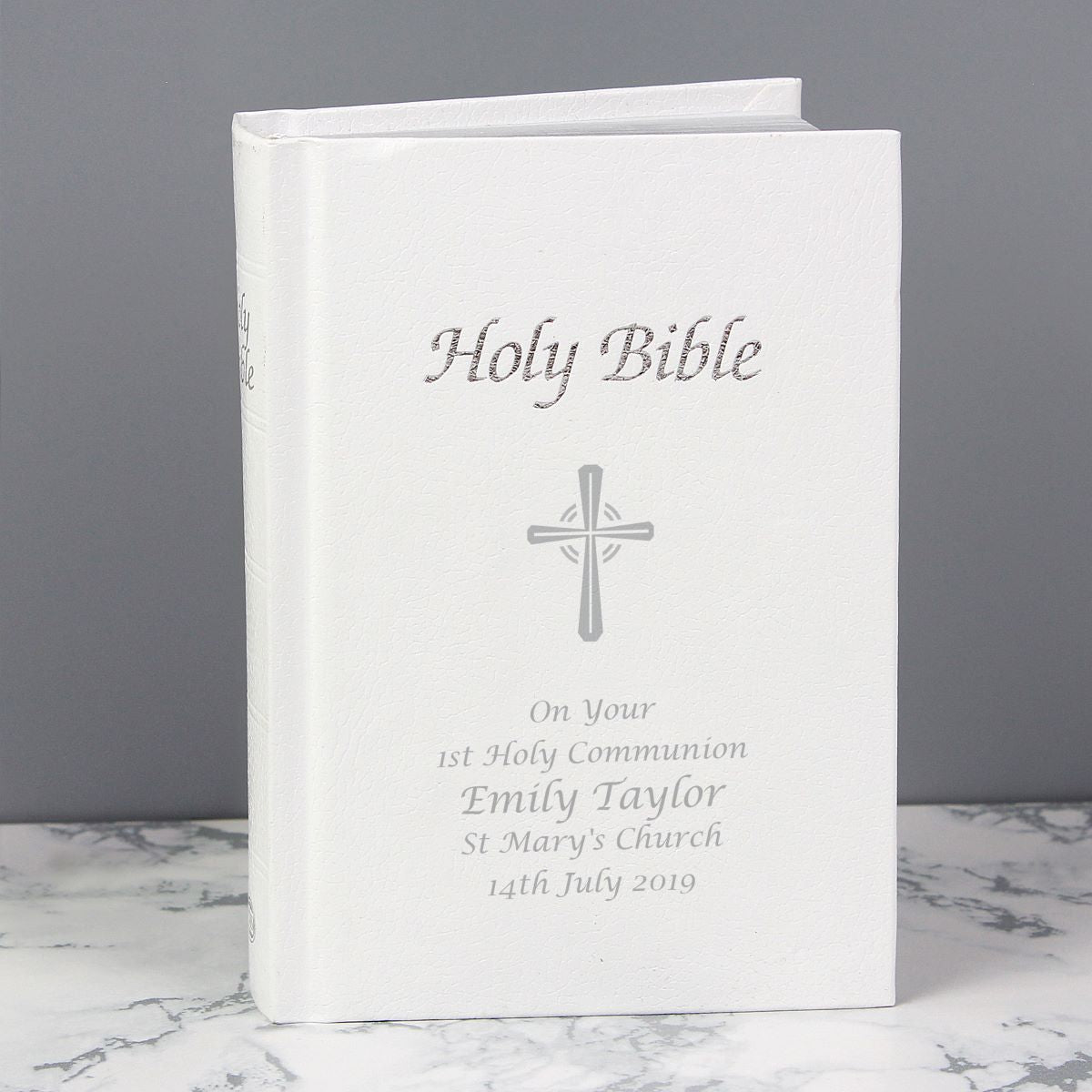 A personalized Holy Bible with a white cover, featuring a silver cross and custom text for a 1st Holy Communion, displayed on a marble surface against a gray background.