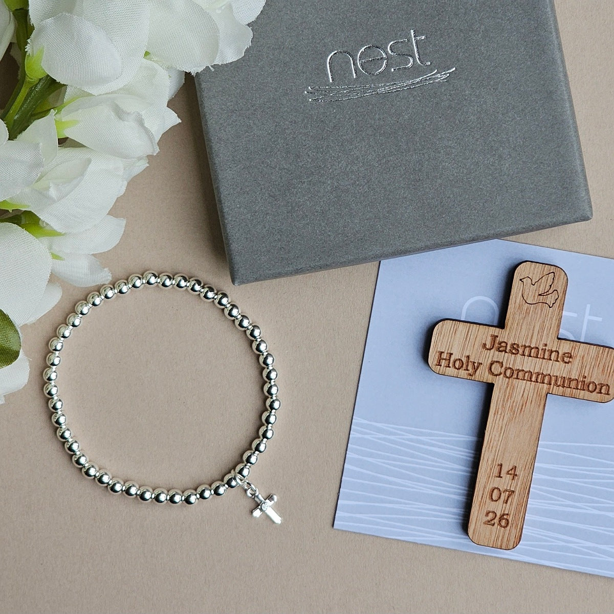 A silver beaded bracelet with a small cross charm is arranged on a beige surface beside a gray Nest gift box, white flowers, and a wooden Holy Communion cross engraved with the name Jasmine and a date.