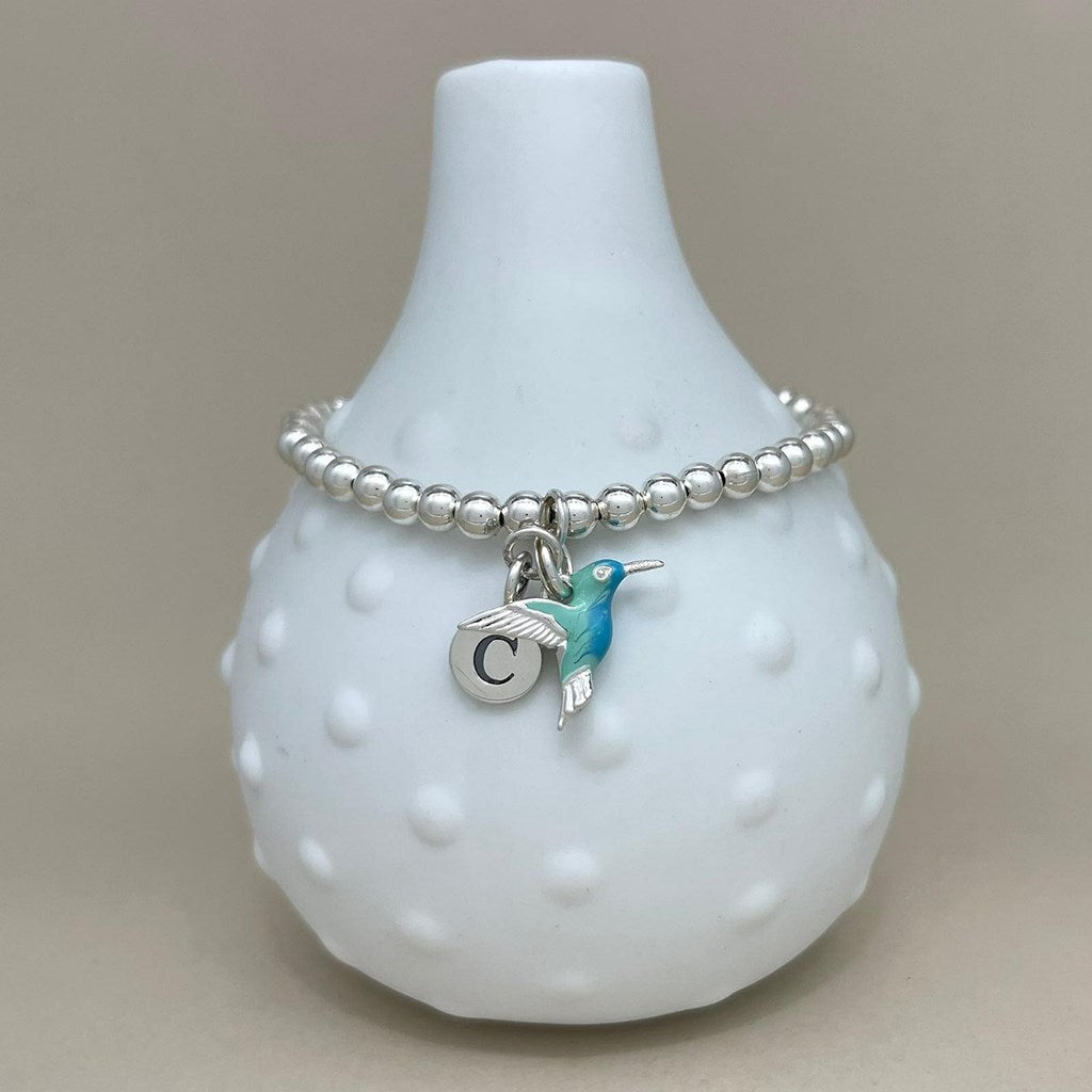 A silver beaded bracelet with a hummingbird charm and a letter 'C' initial charm, displayed on a white textured vase against a neutral beige background.