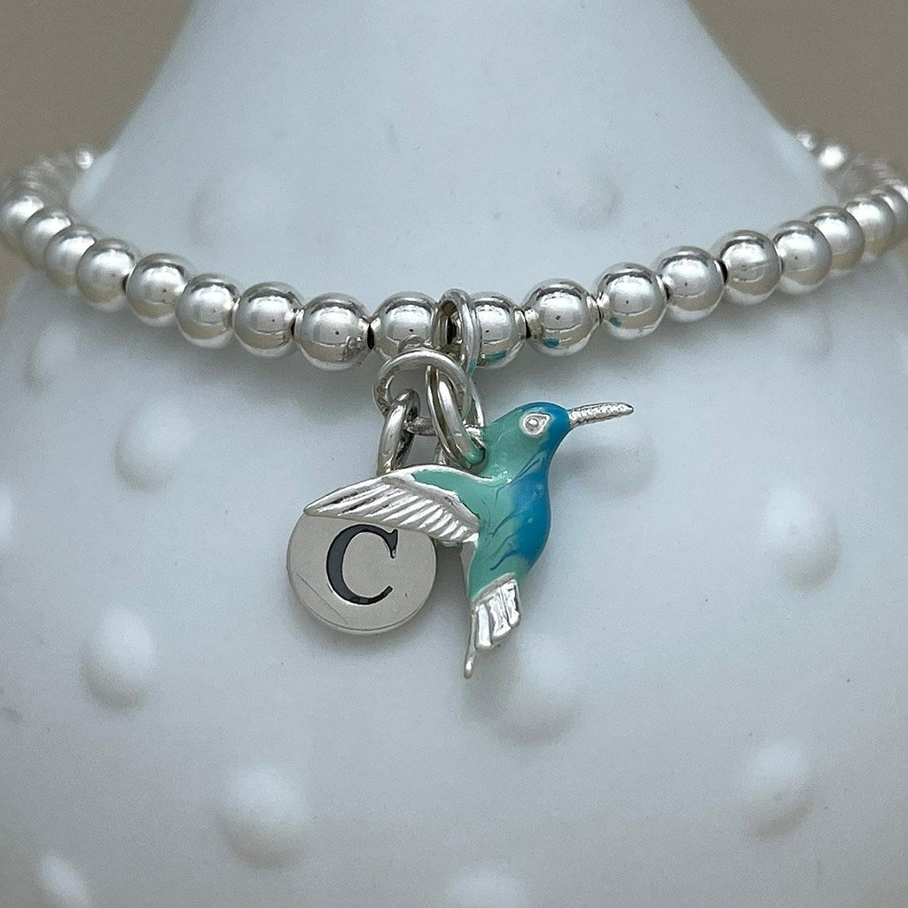 A silver beaded bracelet with a hummingbird charm and a round initial 'C' charm, displayed on a white textured jewelry stand.