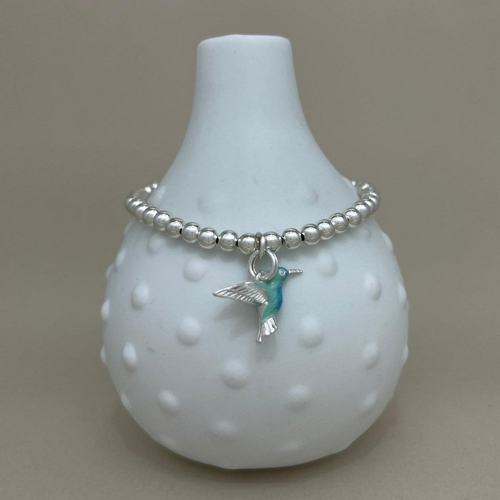 A silver beaded bracelet with a hummingbird charm, displayed on a white textured glass vase against a neutral beige background.