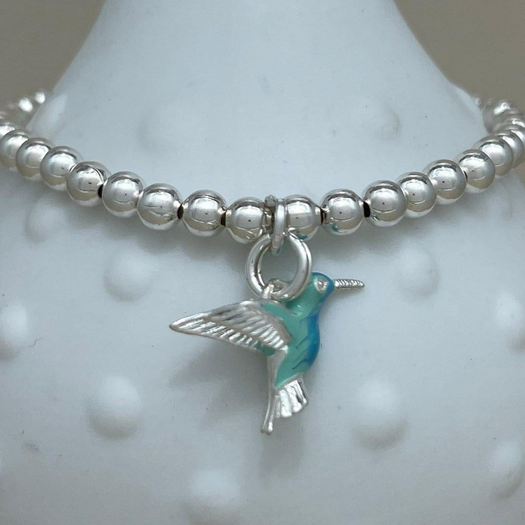 A silver beaded bracelet with a hummingbird charm, displayed on a white textured bust. The bracelet features polished silver beads and a silver-toned hummingbird charm with turquoise blue enamel detailing.