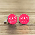 A pair of round silver cufflinks with bright pink faces, featuring white initials with a heart in the middle and a date underneath.