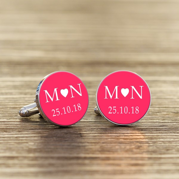 A pair of round silver cufflinks with bright pink faces, featuring white initials with a heart in the middle and a date underneath.