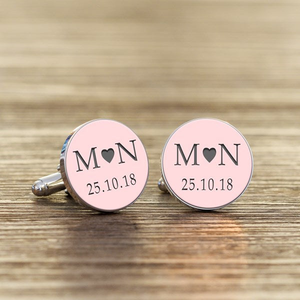 A pair of round silver cufflinks with light  pink faces, featuring grey initials with a heart in the middle and a date underneath.