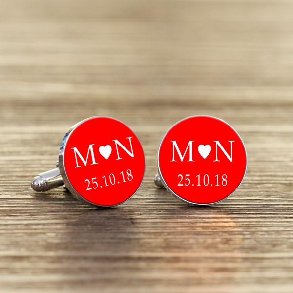 A pair of round silver cufflinks with red faces, featuring white initials with a heart in the middle and a date underneath.