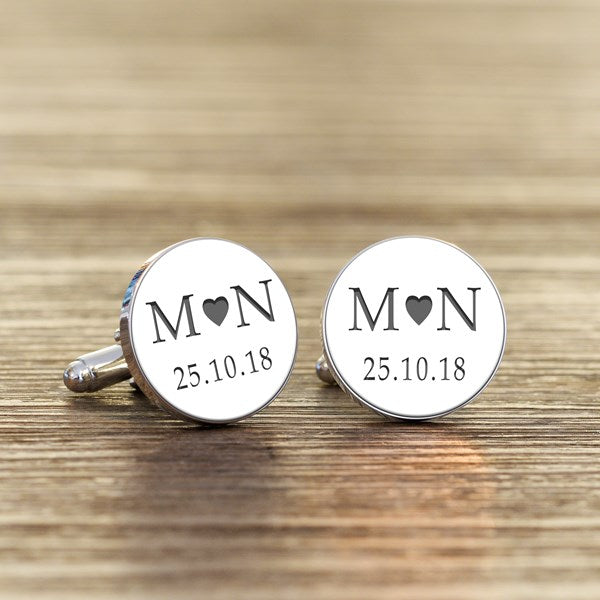 A pair of round silver cufflinks with white faces, featuring grey initials with a heart in the middle and a date underneath.