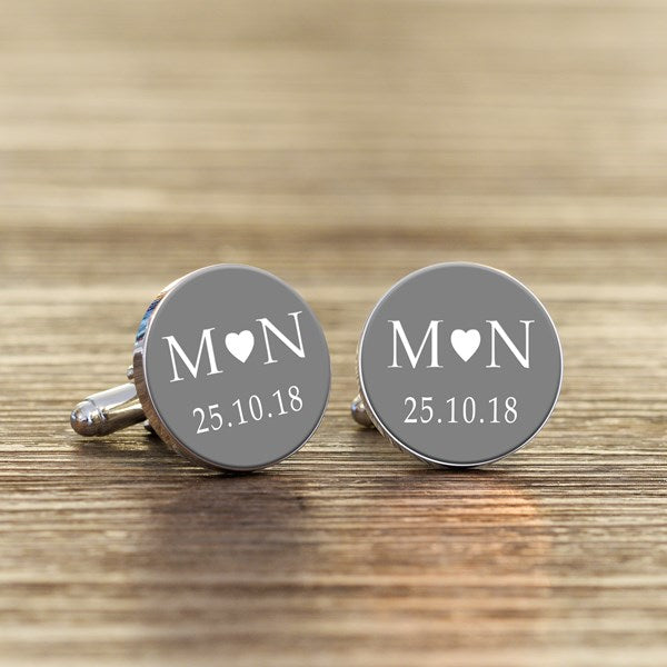 A pair of round silver cufflinks with grey faces, featuring white initials with a heart in the middle and a date underneath.