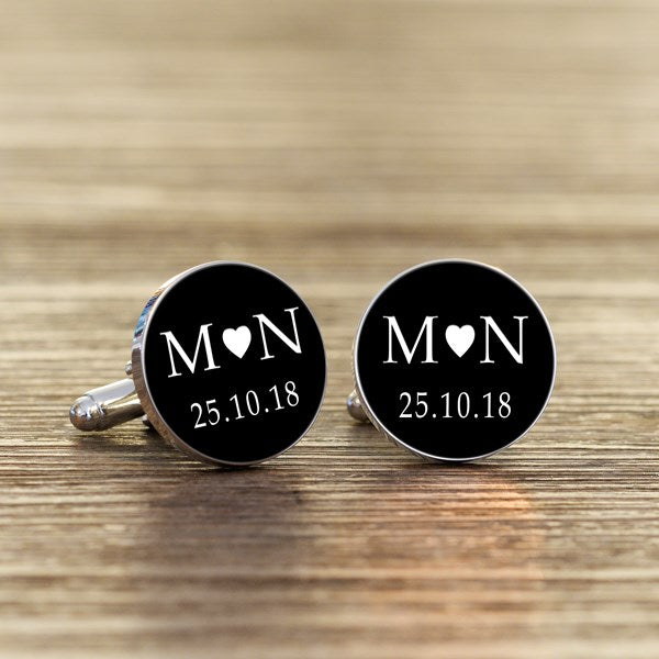 A pair of round silver cufflinks with black faces, featuring white initials with a heart in the middle and a date underneath.