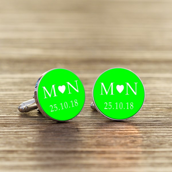 A pair of round silver cufflinks with bright green faces, featuring white initials with a heart in the middle and a date underneath.