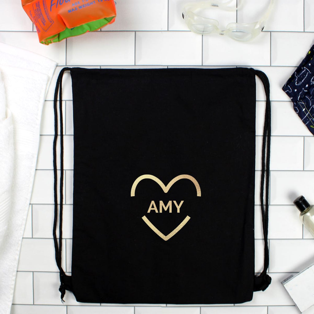 A black drawstring gym bag with a gold heart outline and the name 'AMY' printed on the front, laid flat on a white tiled surface with small lifestyle props around it.