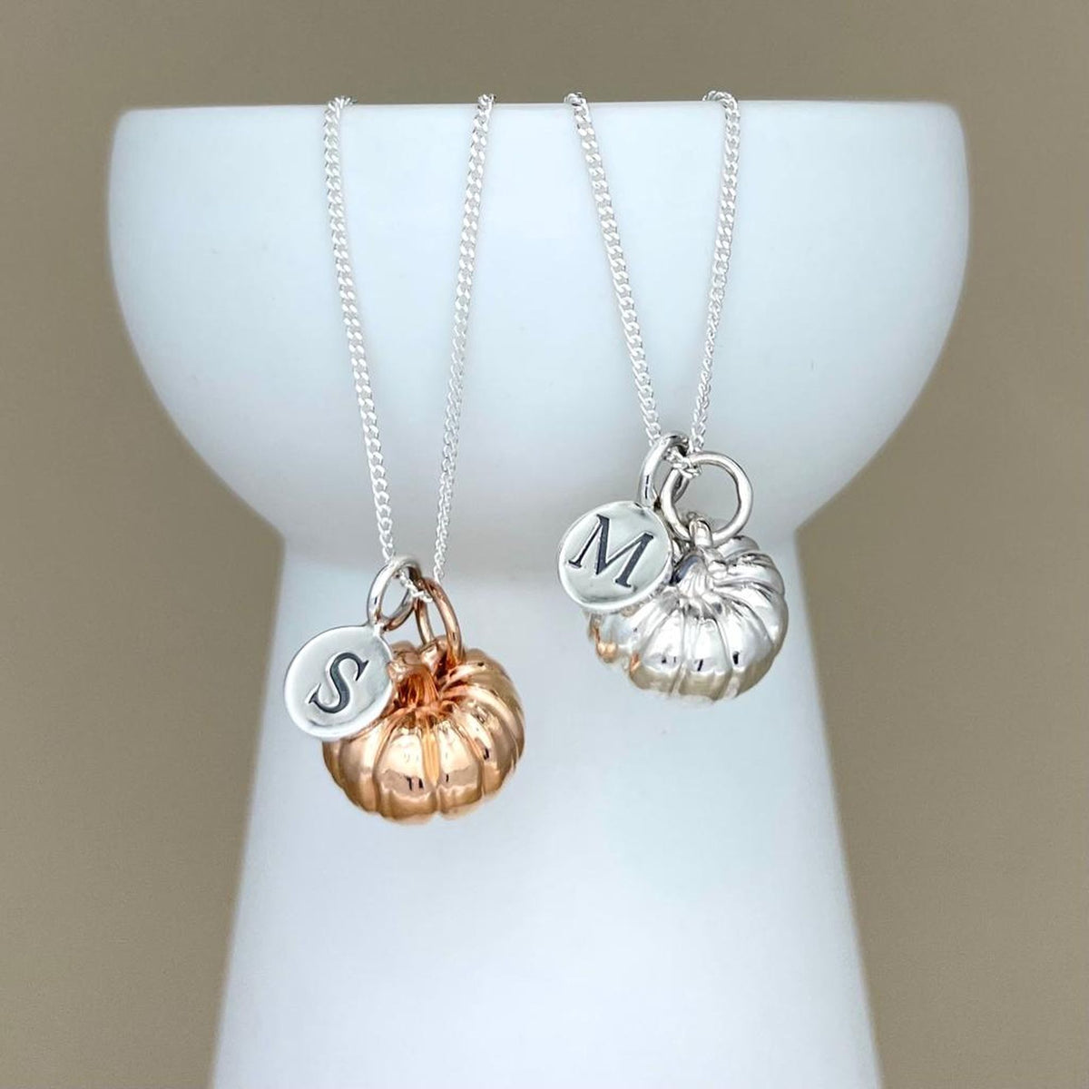 Two silver necklaces with initial charms and pumpkin charms on a white stand against a beige background