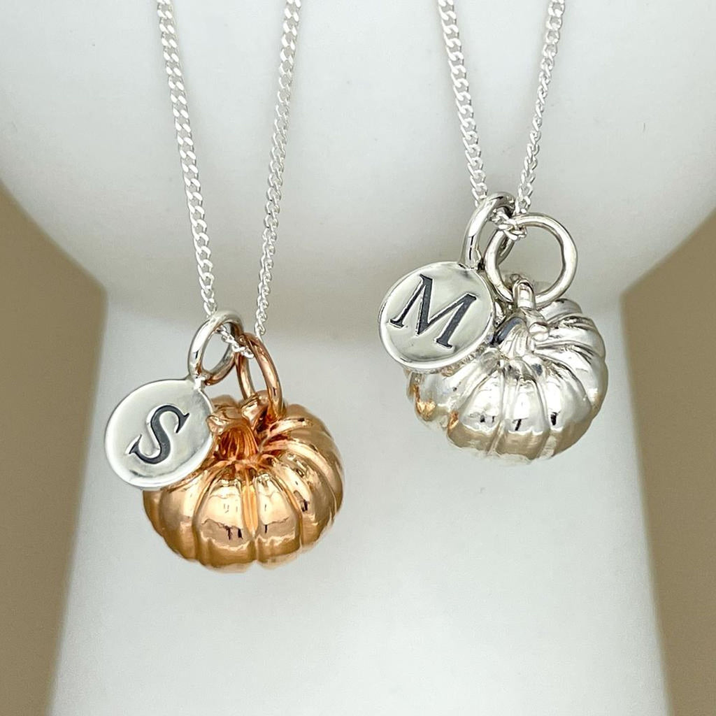 Two silver pendant necklaces displayed on a white jewelry bust. Each necklace has a silver chain, a small round silver initial charm engraved with a letter (S and M), and a pumpkin-shaped charm in a different metal finish: one rose gold and one silver.