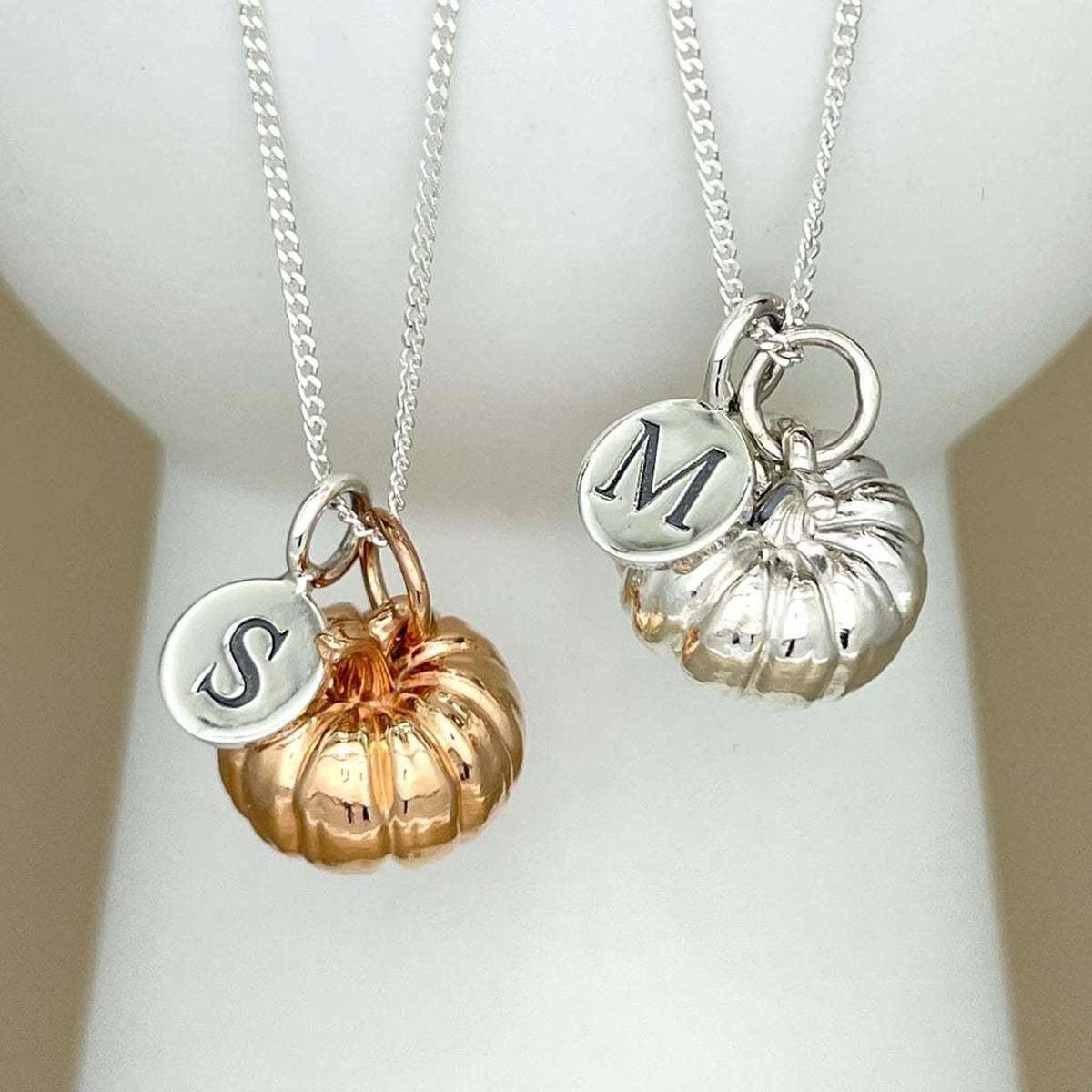 Two silver pendant necklaces displayed on a white jewelry bust. Each necklace has a silver chain, a small round silver initial charm engraved with a letter (S and M), and a pumpkin-shaped charm in a different metal finish: one rose gold and one silver.