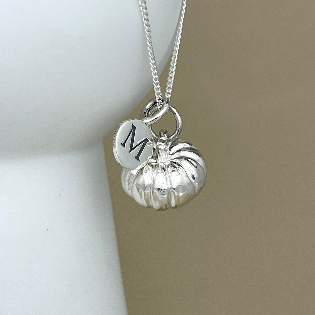 A silver necklace featuring a small pumpkin charm and a round disc charm engraved with the letter 'M', displayed on a neutral beige and white background.
