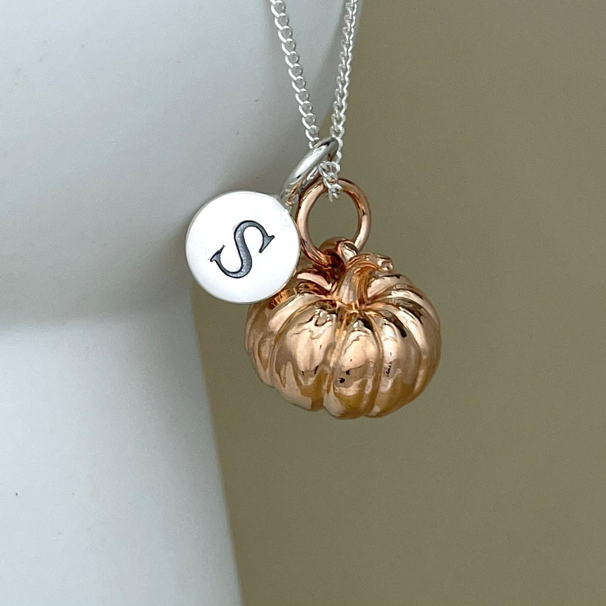 A silver chain necklace with a polished rose gold pumpkin charm and a round silver disc charm engraved with a black monogram, shown against a neutral beige background.