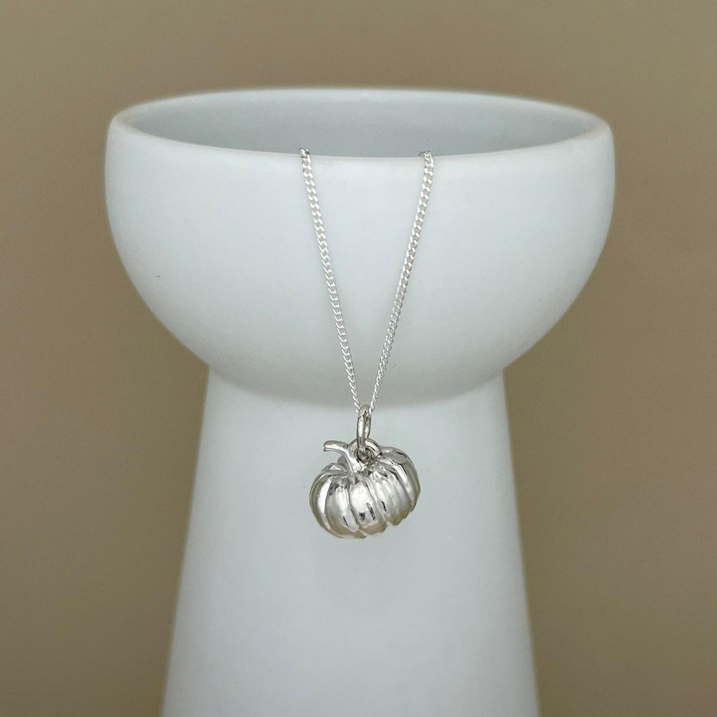 A silver pumpkin pendant necklace on a delicate silver chain, displayed draped over a white ceramic vase against a solid beige background.