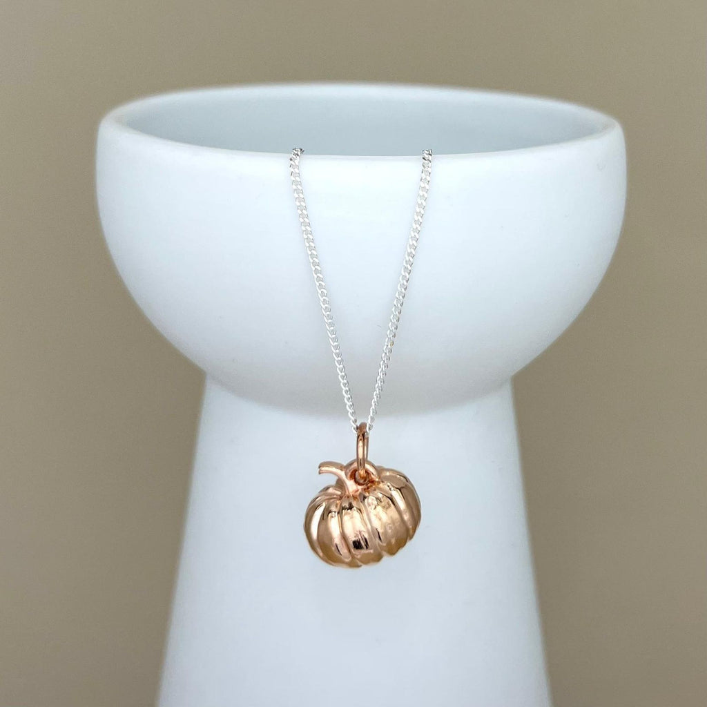 A rose gold pumpkin pendant necklace on a delicate silver chain, displayed draped over a white ceramic pedestal against a warm beige background.