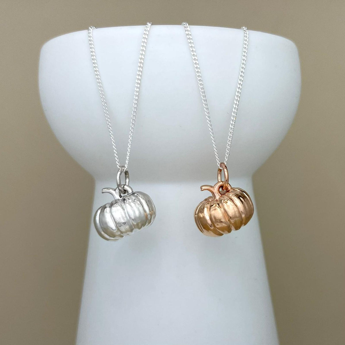 Two pumpkin-shaped pendant necklaces hanging from a white display bust, one in polished silver and one in polished rose gold, against a plain beige background.